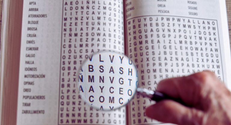 Main tenant une loupe sur une grille de mots fléchés pour chercher la solution du mot âpre au gain en 6 lettres